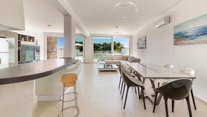 A modern kitchen with a central island, a dining area with a glass table, and a view of the outdoors. - 1934 Villa Punta Ristola (Castrignano del Capo)