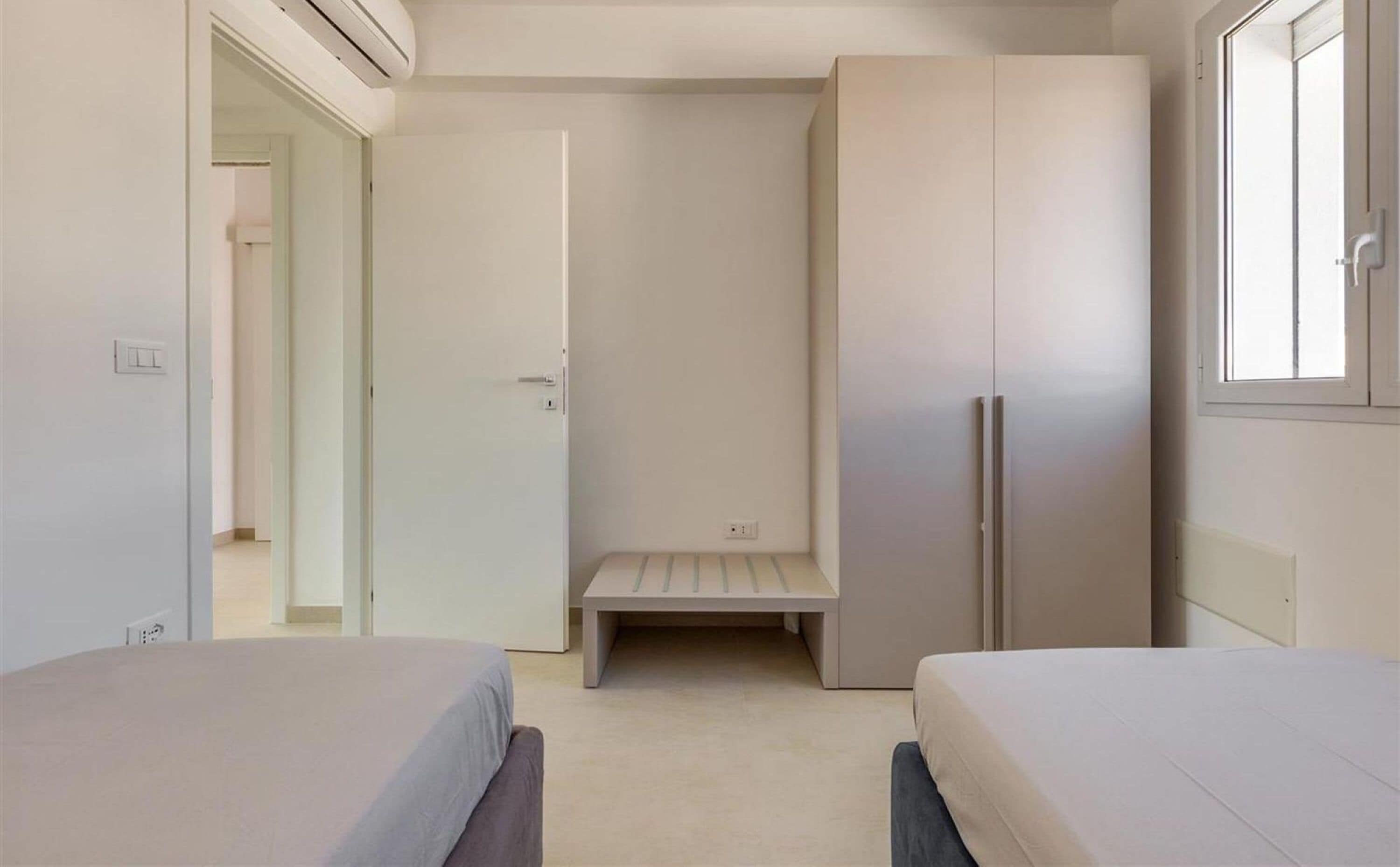 A bedroom with two beds, a wardrobe, a bench, and a window.