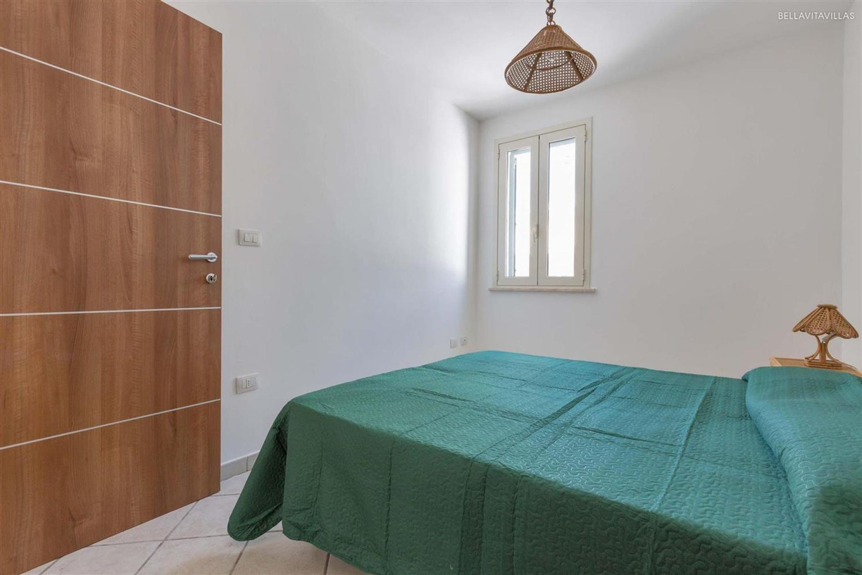 A bedroom with a wooden wardrobe, a single bed with a green bedspread, a window with blinds, and a hanging light fixture.