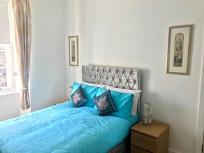 A neatly made bed with a patterned headboard, a blue bedspread, and decorative pillows. There is a nightstand with a vase and a framed picture on the wall. - Stunning 3-bed Apartment in Edinburgh (Edinburgh)