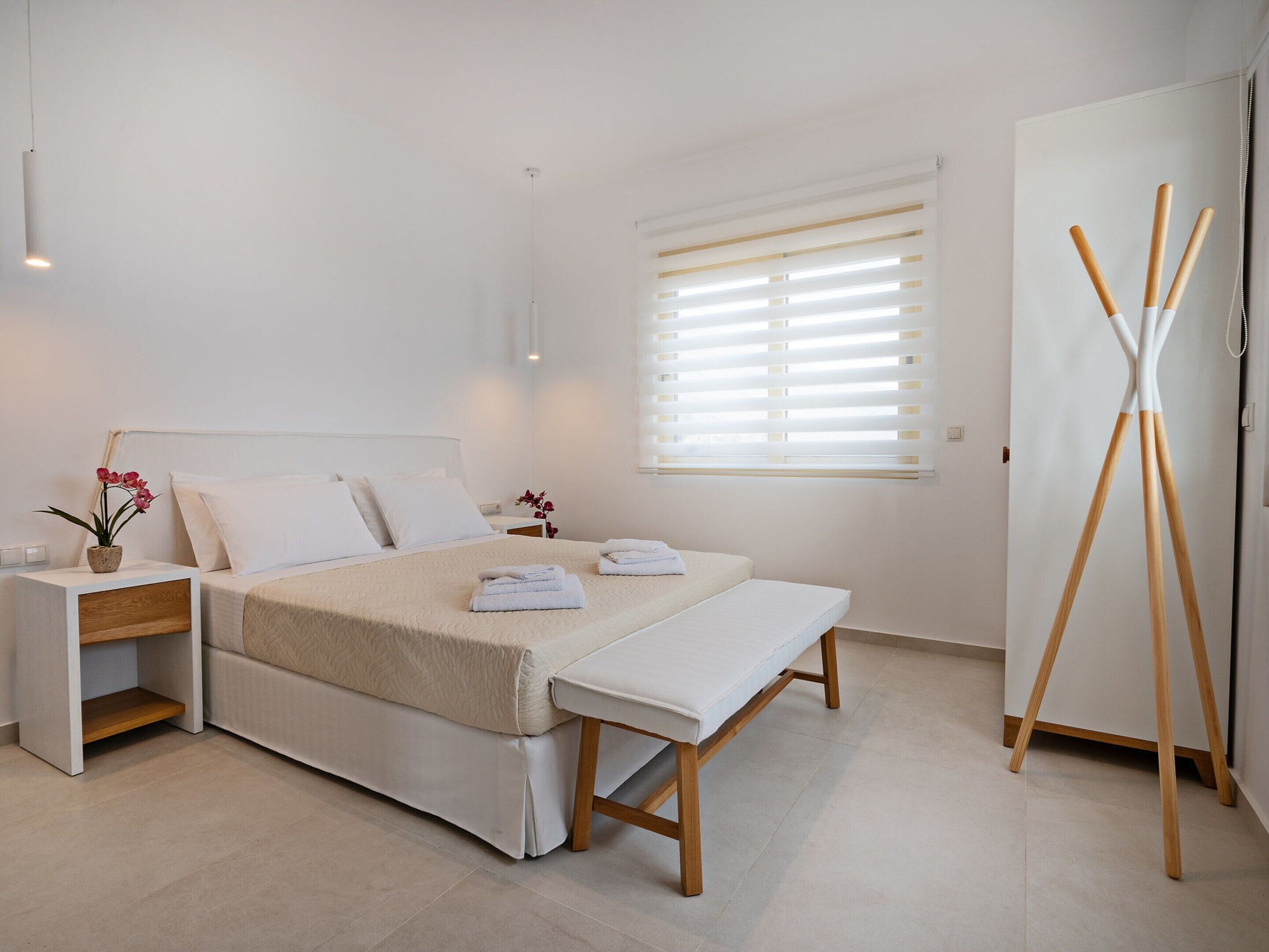 A minimalist bedroom with a bed, bedside tables, a bench, a window with blinds, and a wooden stand.