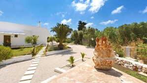 Un bâtiment blanc avec un escalier menant à une porte, entouré de verdure et d’une grande formation rocheuse.