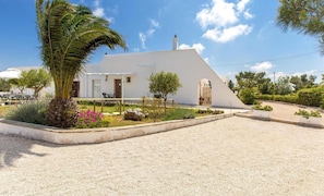 A modern building with a white facade, a small patio with a table and chairs, and a landscaped garden with a palm tree and shrubs.