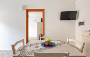 Uma área de refeições com uma mesa posta para dois, uma fruteira sobre a mesa, uma televisão fixada na parede e um armário de madeira ao lado.