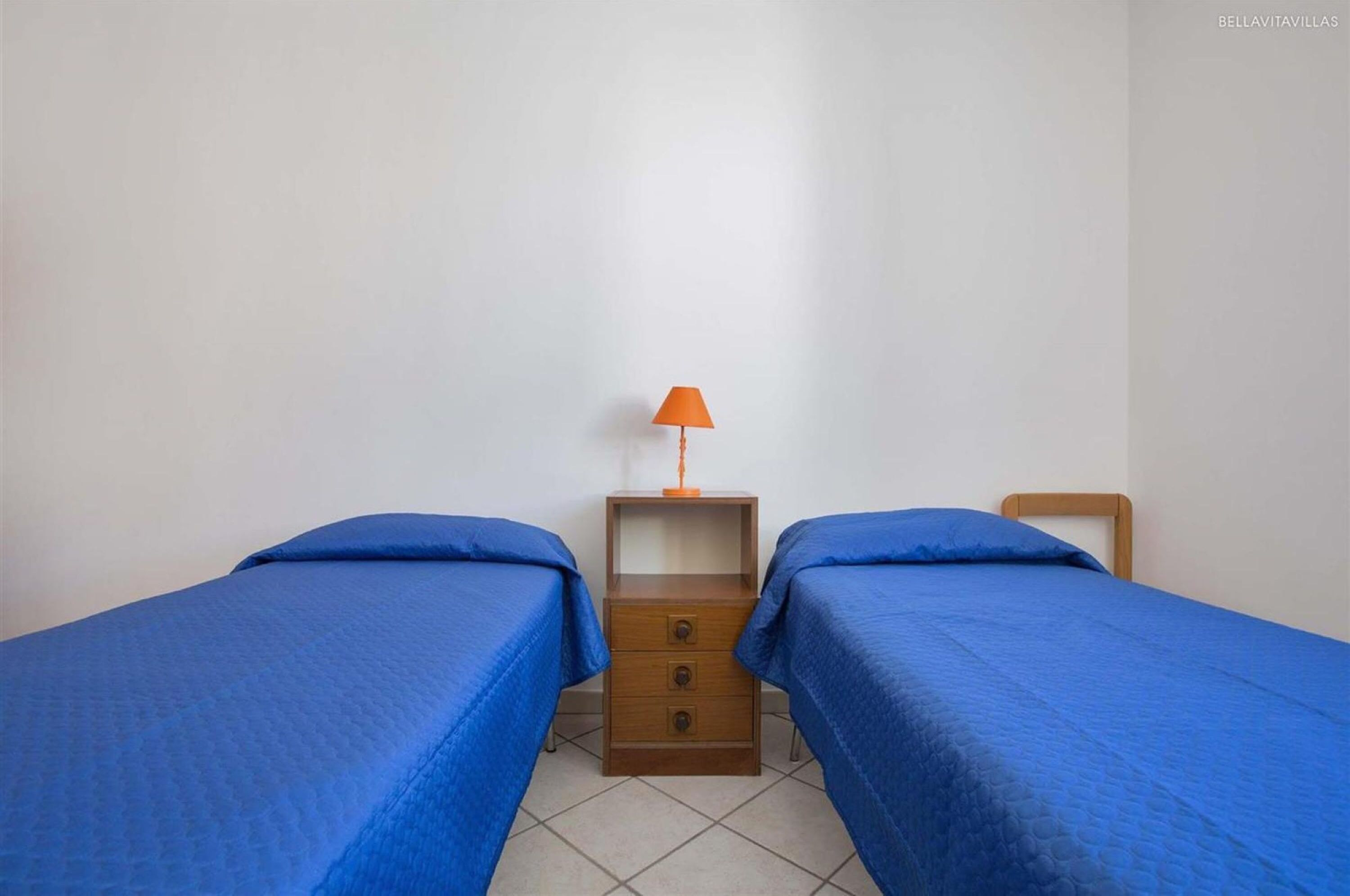 A room with two single beds, each with a blue bedspread, a wooden nightstand with an orange lamp, and a wooden headboard.