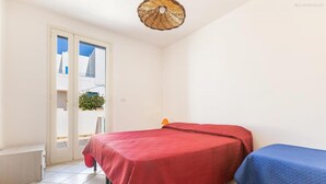 Une pièce avec un lit recouvert d’un tissu rouge, un plus petit lit avec une couverture bleue, un luminaire suspendu en osier au plafond et une porte donnant sur l’extérieur.