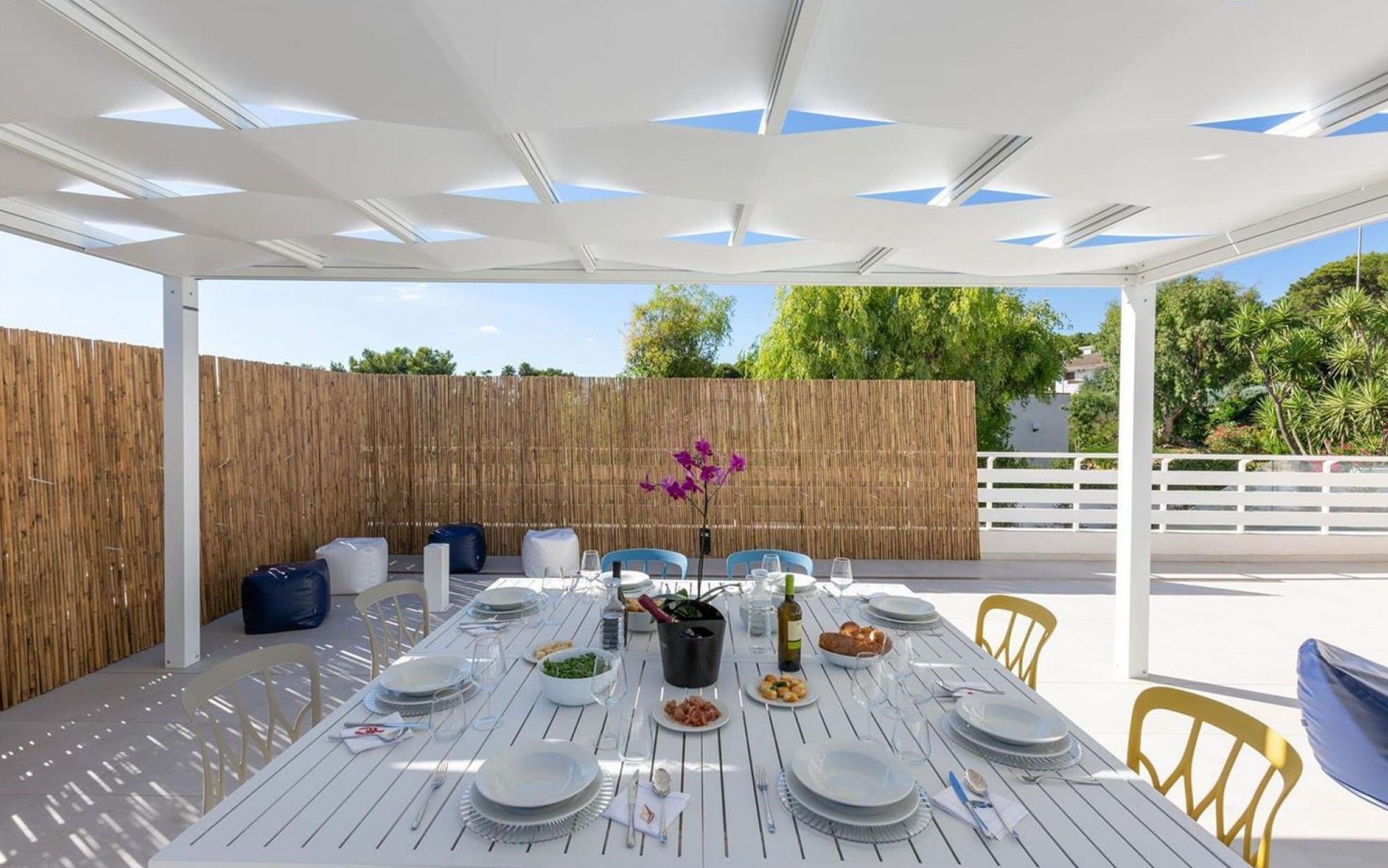 Un'area pranzo all'aperto con un tavolo bianco imbandito per quattro persone, circondato da sedie gialle, sotto un pergolato bianco con dettagli blu.