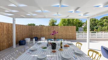 Un'area pranzo all'aperto con un tavolo bianco imbandito per quattro persone, circondato da sedie gialle, sotto un pergolato bianco con dettagli blu.