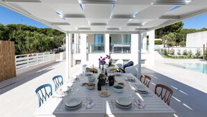 Un área de comedor al aire libre con una mesa blanca dispuesta para una comida, rodeada de sillas y con vistas a una piscina.