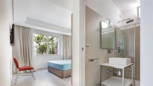 Una habitación de hotel con cama, una silla roja, una ventana con cortinas, un baño con lavabo y espejo, y ducha.