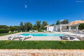 Une piscine avec des chaises longues, un espace de détente couvert et un mur en pierre.
