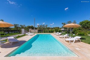 Une piscine rectangulaire à l’eau d’un bleu cristallin, entourée de chaises longues blanches et de parasols beige.