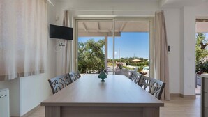 A dining area with a long table and chairs, a flat-screen TV mounted on the wall, and a view of the outdoors. - 2390 Villa Eva by Perle di Puglia (San Vito dei Normanni)