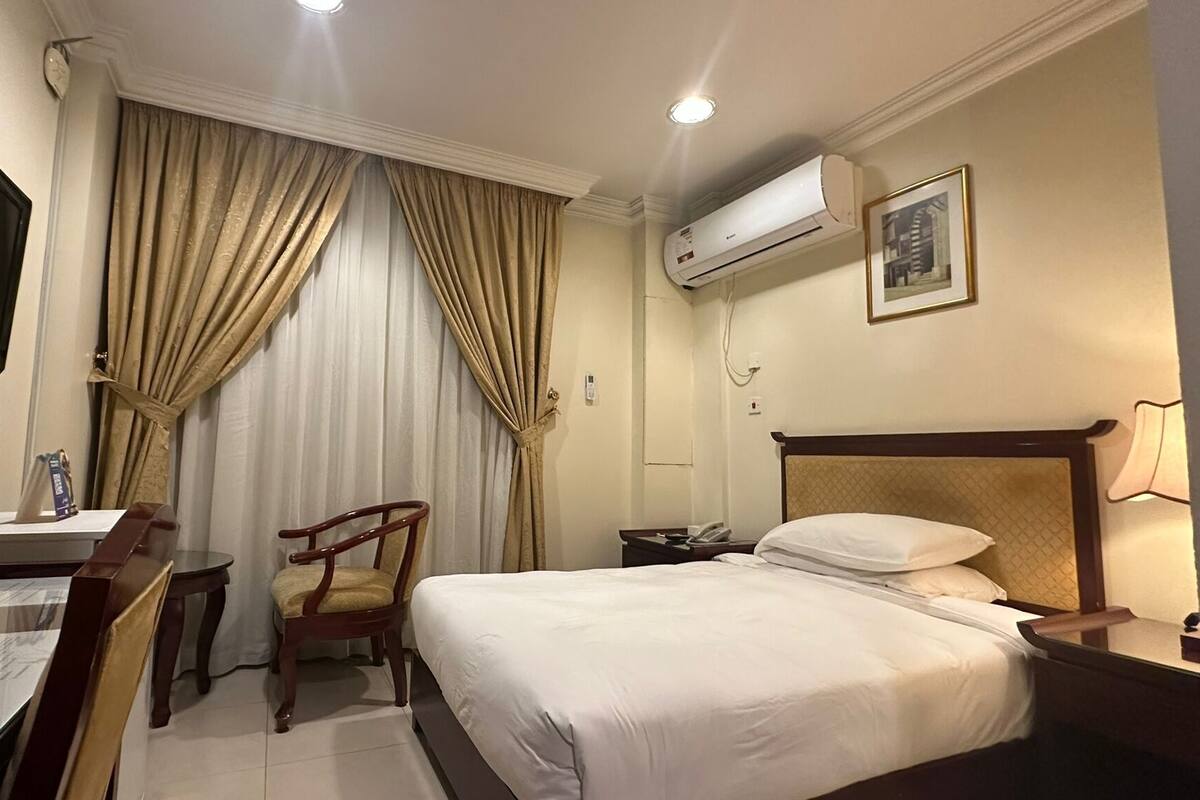Habitació d'hotel amb un llit, un escriptori amb una cadira, una televisió, un aparell d'aire condicionat i una taula de nit amb una làmpada.