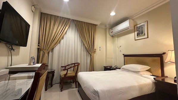 Habitación de hotel con cama, escritorio con silla, televisor, equipo de aire acondicionado y mesita de noche con lámpara.