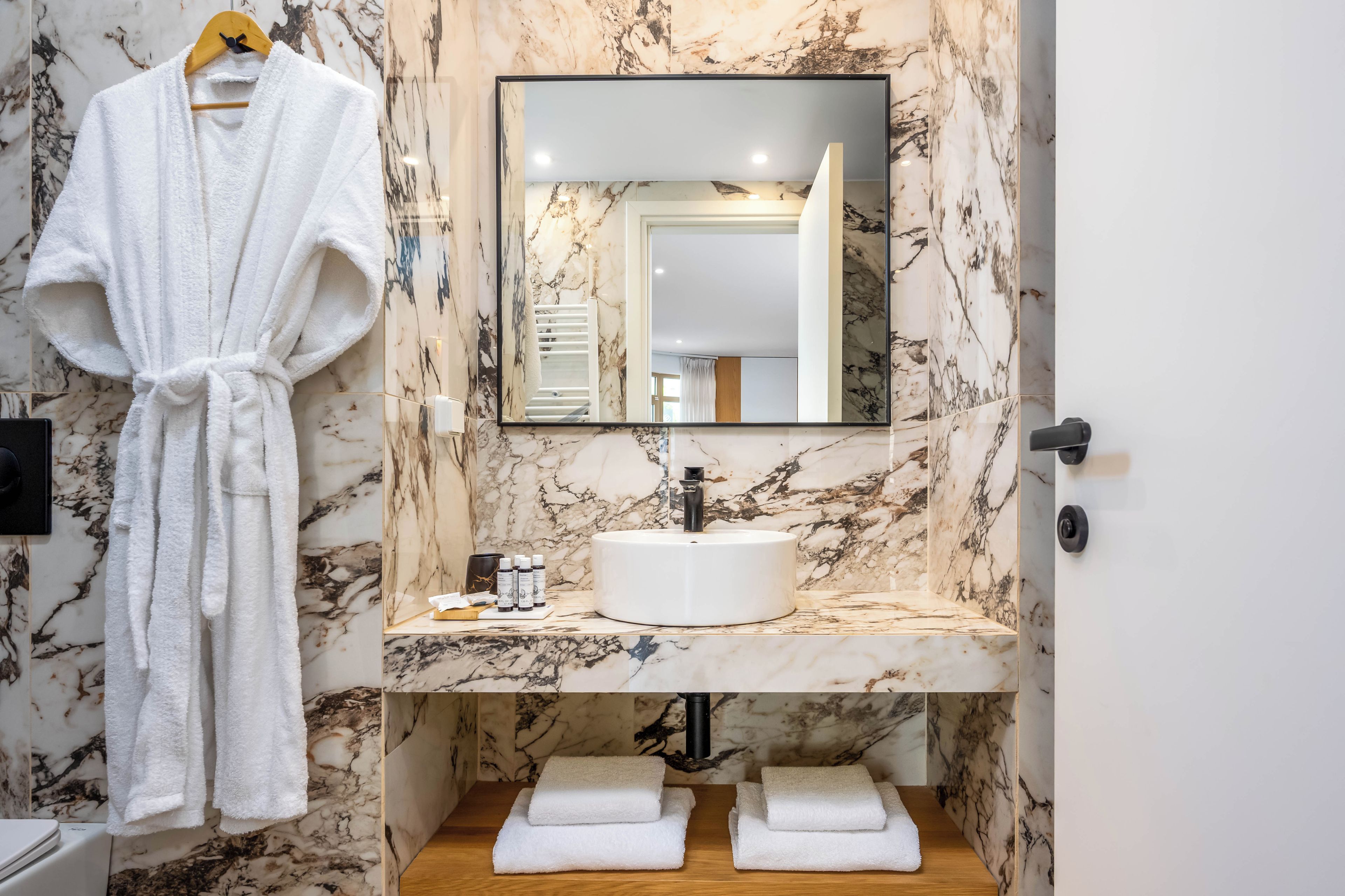 Une salle de bain avec un mur en marbre, un lavabo blanc, un miroir et une robe suspendue à un porte-serviettes.