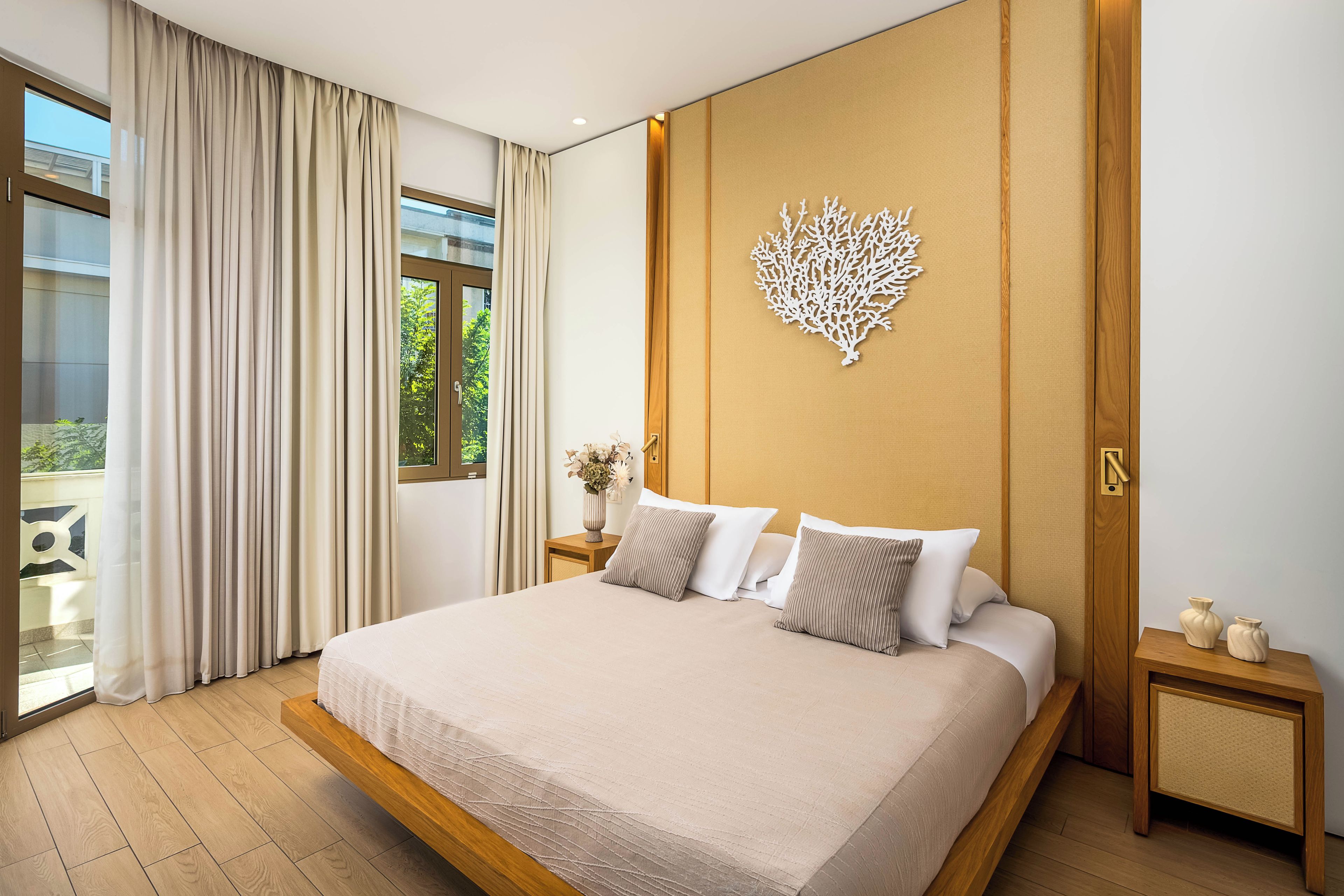 A modern bedroom with a large bed, a wooden headboard, a decorative wall piece, and a view of greenery outside.