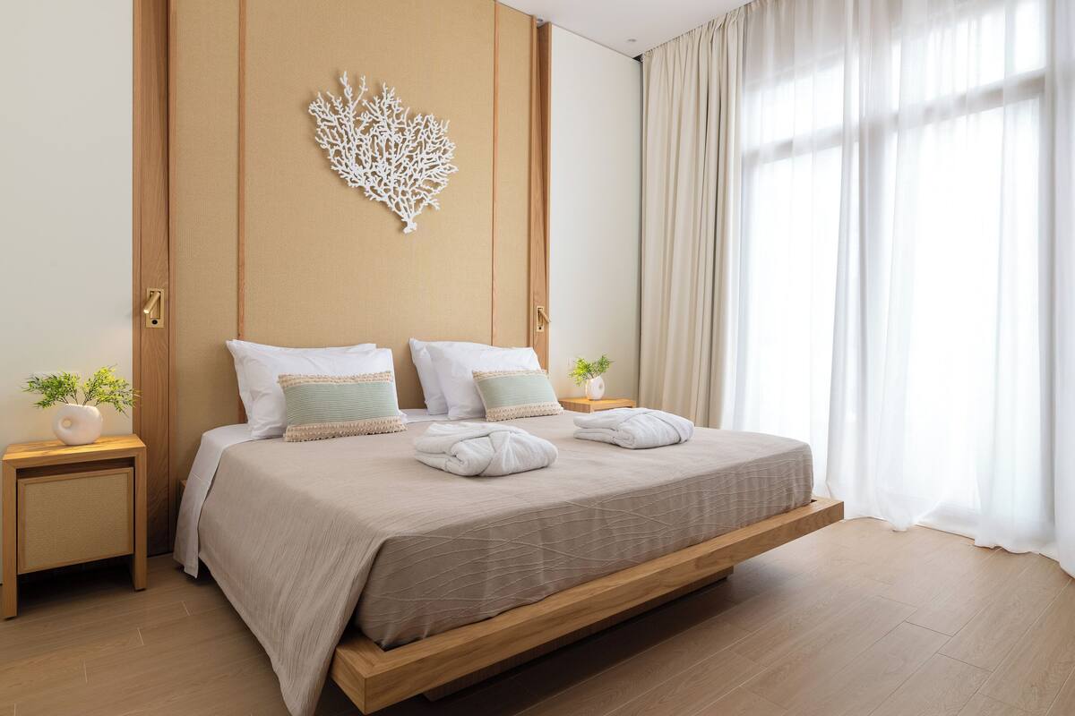 A modern bedroom with a bed, nightstands, a decorative wall piece, and a view through sheer curtains.