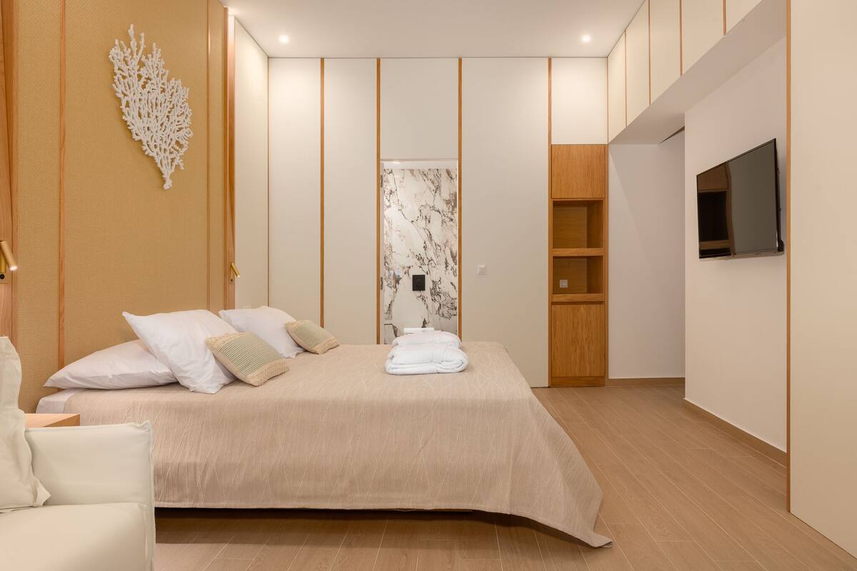 A modern bedroom with a large bed, a wooden wardrobe, a TV mounted on the wall, and a marble-patterned door.