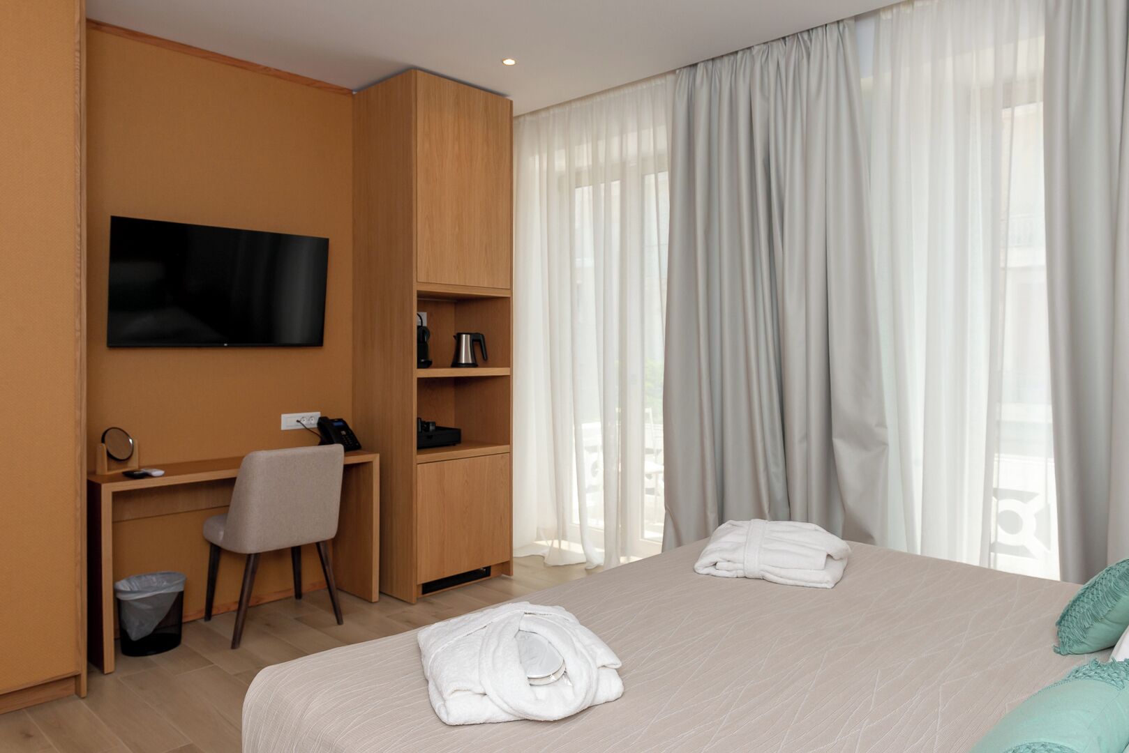 Habitación de hotel con cama, escritorio con silla, televisor montado en la pared y ventana con cortinas.