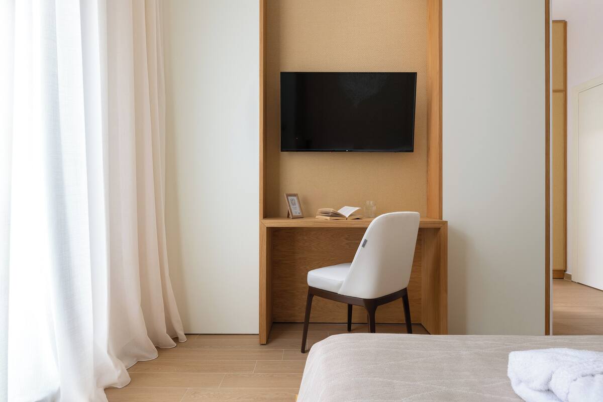 A hotel room with a bed, a desk with a chair, a flat-screen TV mounted on the wall, and a window with curtains.