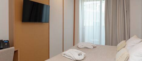 A hotel room with a bed, a TV mounted on the wall, a desk with a telephone, and a window with curtains.