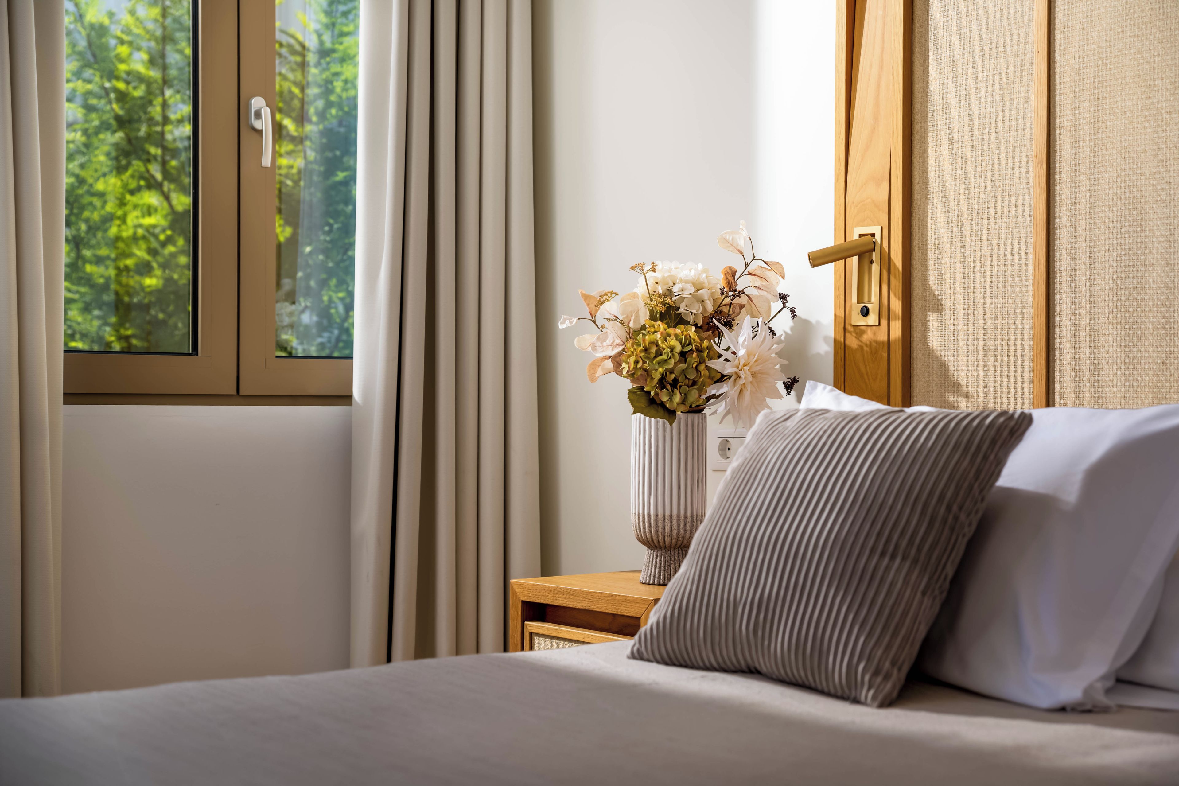 A bedroom with a bed, a nightstand, a vase with flowers, and a window with curtains.