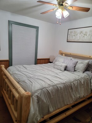 Une chambre à coucher avec un cadre de lit en bois, un ventilateur de plafond, une lampe fixée au mur et une œuvre d’art florale encadrée.