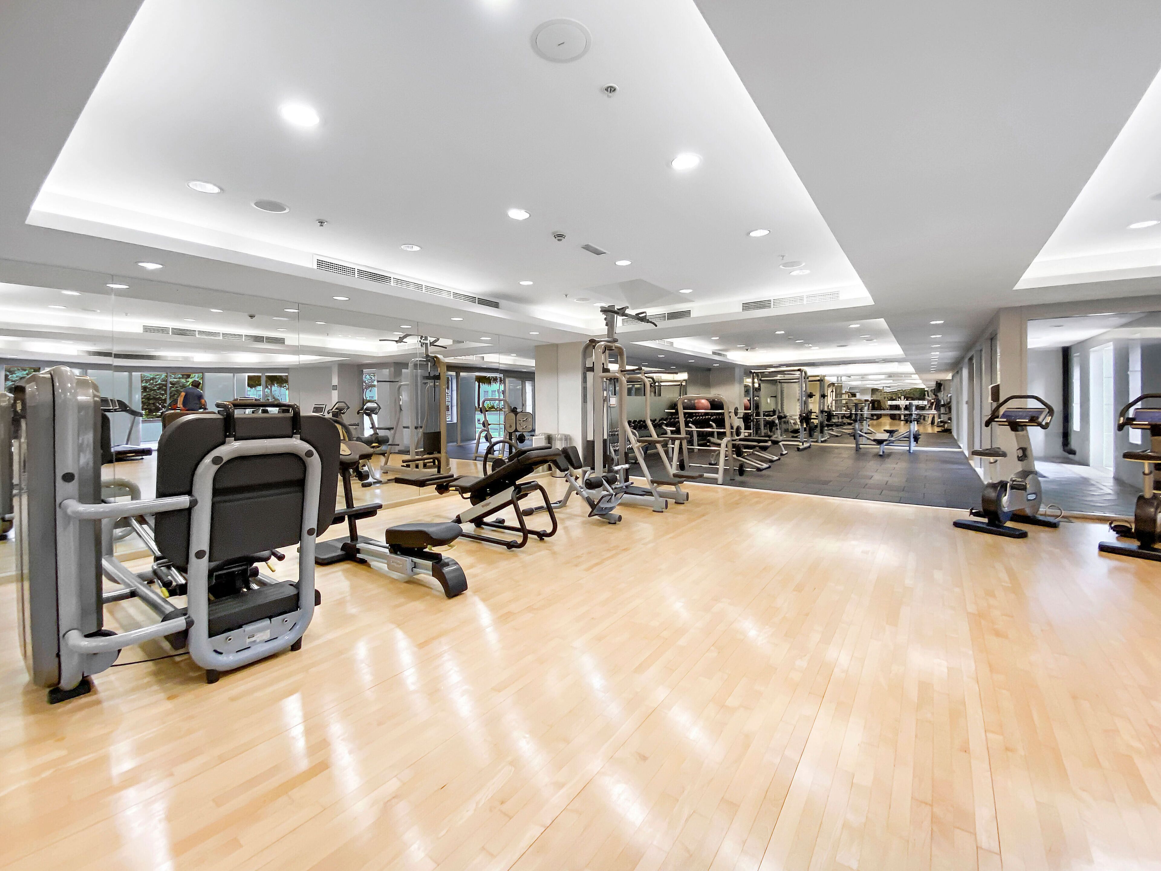 Un gimnasio amplio con diversas máquinas de ejercicio, piso de madera y una gran pared de espejos.