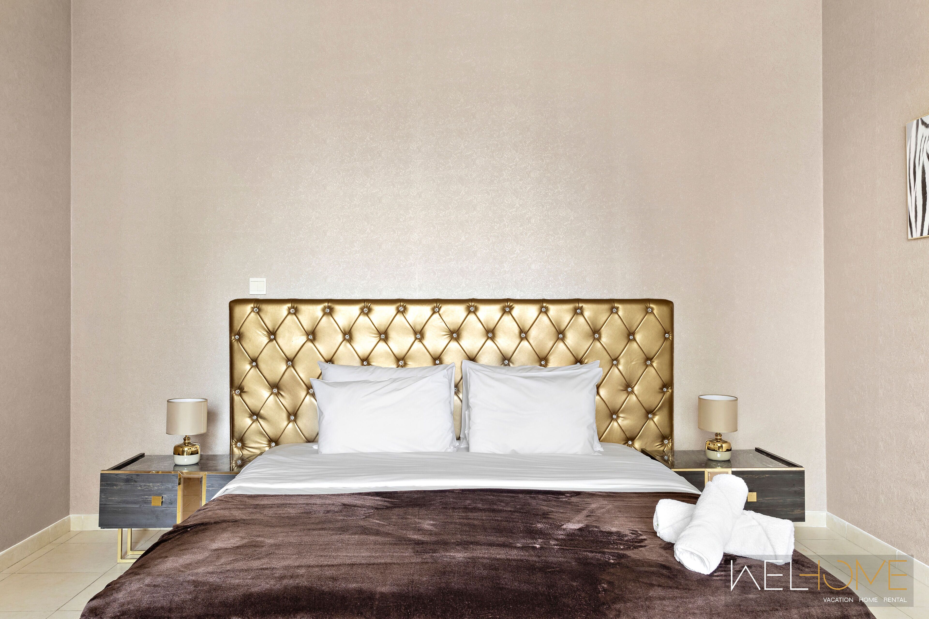 Una habitación de hotel con un cabecero capitonado, una cama con sábanas blancas, dos mesitas de noche con lámparas y una manta marrón sobre la cama.