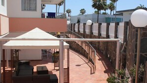 Un espacio de descanso al aire libre cubierto con sofá y sillones, una pérgola y vistas a un edificio residencial con balcón.
