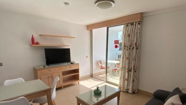 Una sala de estar moderna con un sofá, una mesa de comedor con sillas, un televisor y un ventanal corredizo que da a un área exterior.
