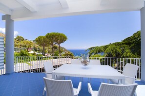 Eine Terrasse mit weißem Tisch und Stühlen, mit Blick auf eine Küstenlandschaft mit Bäumen und Meer.
