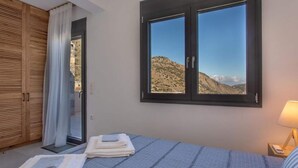 A bedroom with a bed, a window with a view of hills, a wooden wardrobe, and a nightstand with a lamp. - Mesmerising Villa in Plaka Elounda With Pool (Agios Nikolaos)