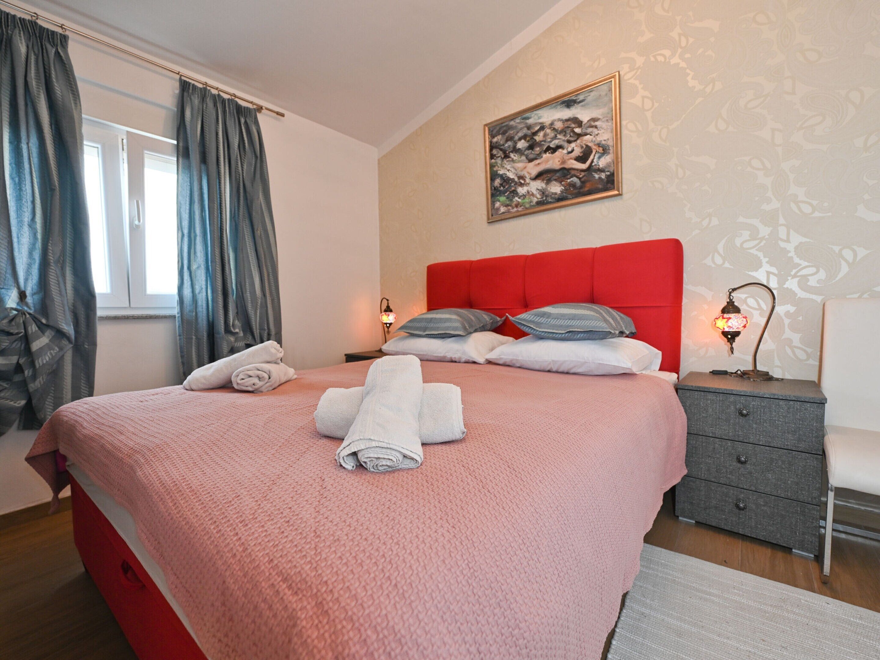 A neatly made bed with a red headboard, two bedside lamps, and a painting above it.