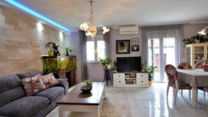 A modern living room with a grey sofa, a coffee table, a TV, and a dining area with floral chairs. - Home With Swimming Pool & Outdoor Kitchen (Nin)