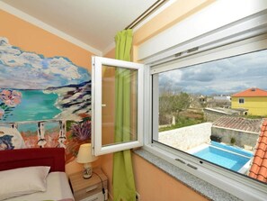 A bedroom with a painting of a coastal scene, a bed, a nightstand with a lamp, and a window overlooking a swimming pool and buildings. - Home With Swimming Pool & Outdoor Kitchen (Nin)
