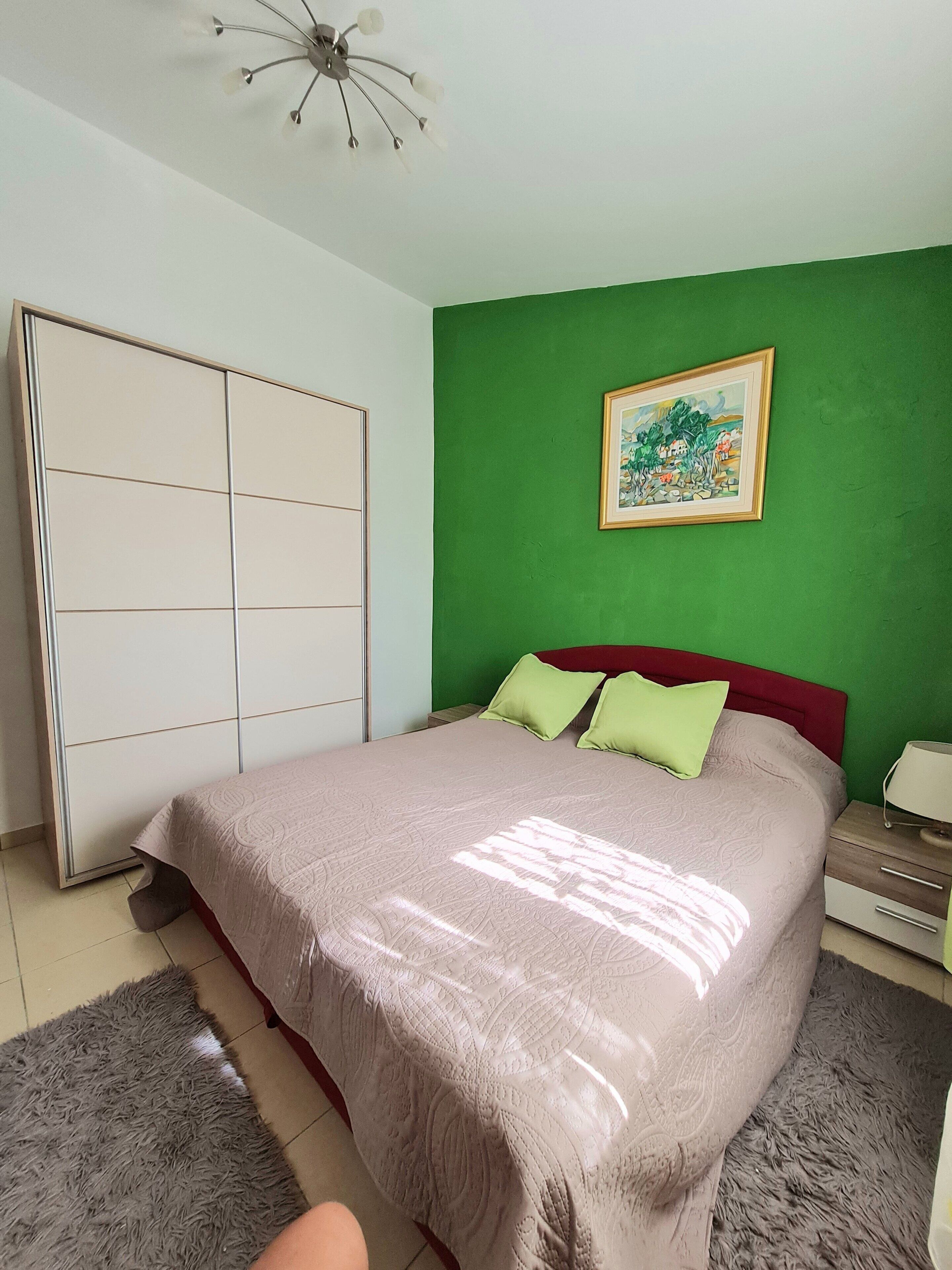 Un dormitorio con cama, paredes verdes, ventilador de techo, armario y un cuadro en la pared.