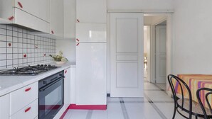 Une cuisine avec des meubles blancs, une cuisinière à gaz et un dosseret carrelé. On aperçoit une petite plante sur le comptoir et une chaise recouverte d’une nappe à rayures.