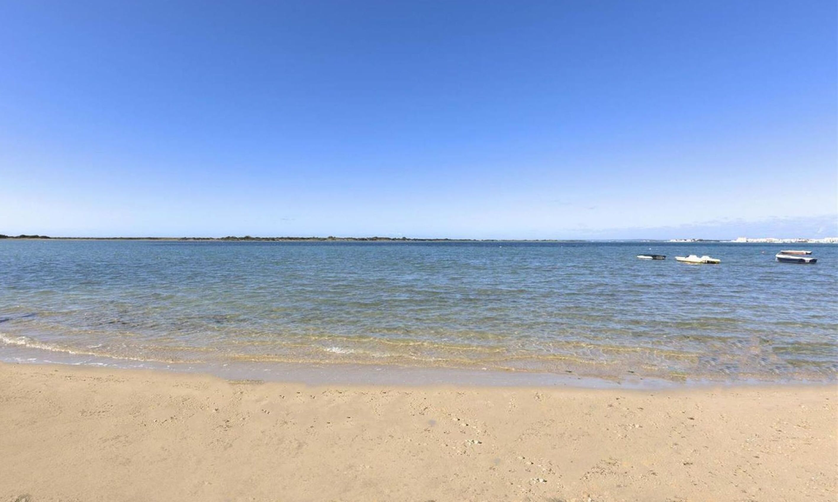 A beach with clear water, a sandy shore, and a few boats in the distance.