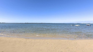 Une plage à l’eau claire, avec un rivage sablonneux et quelques bateaux visibles au loin.