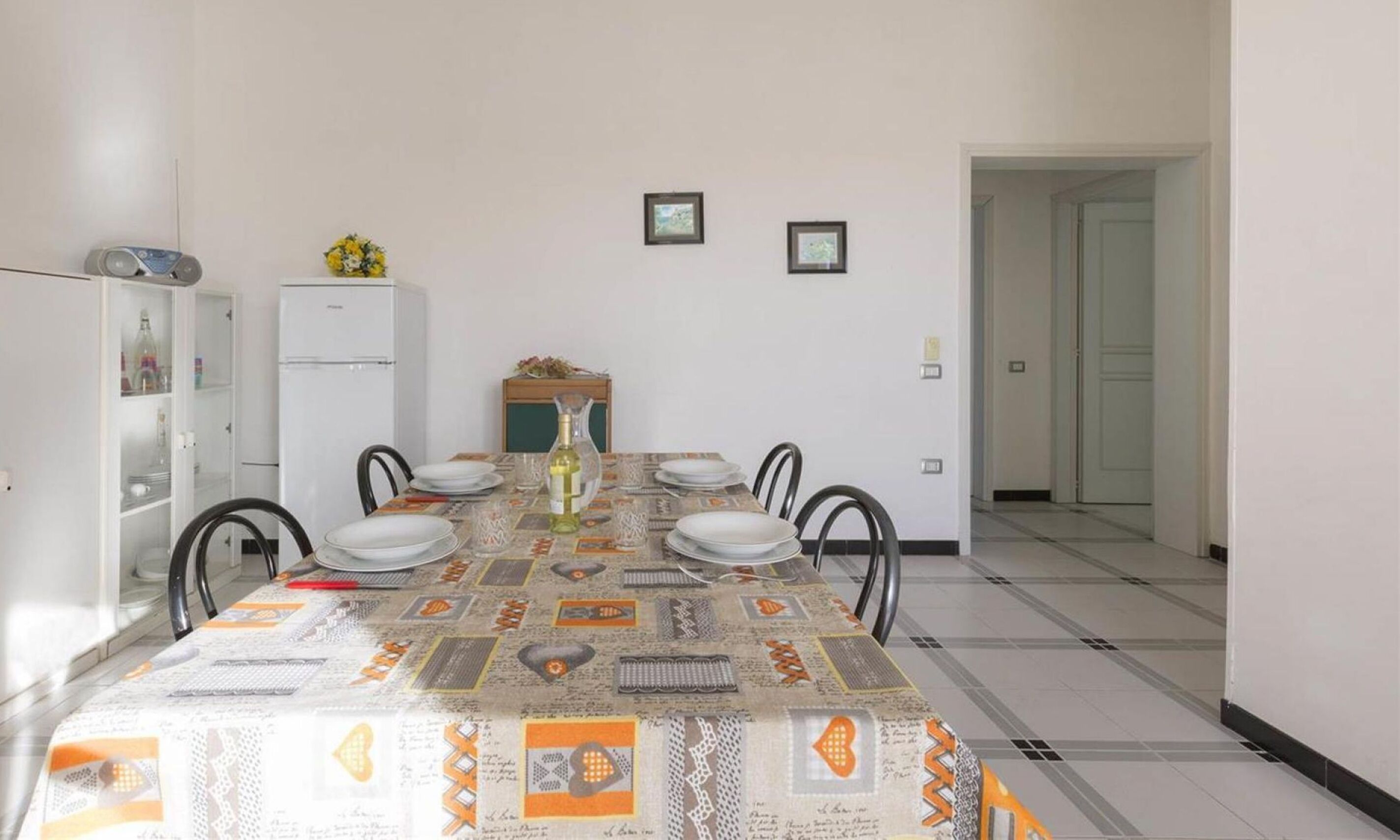 A dining area with a patterned tablecloth, white walls, and a refrigerator.