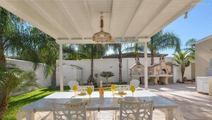 An outdoor patio with a table set for two, surrounded by white chairs, under a white pergola.