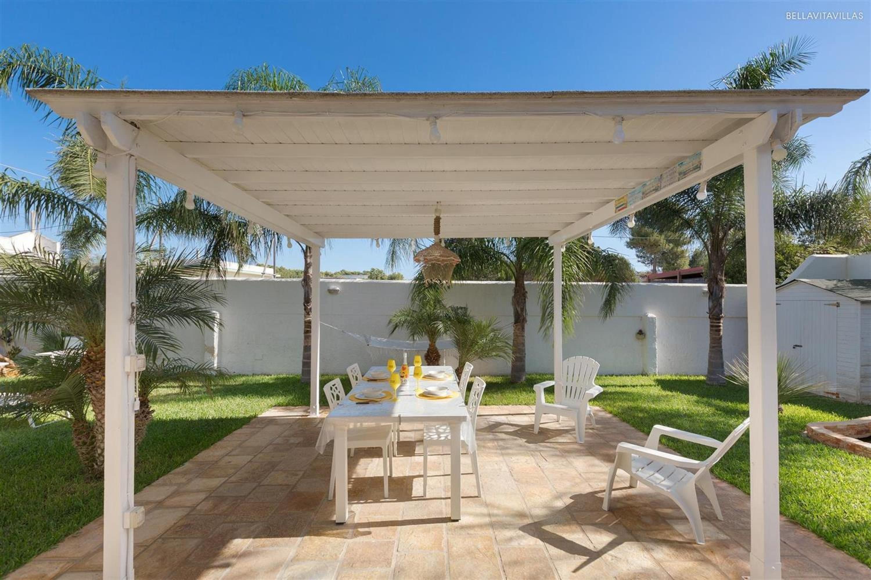 A covered patio with a table set for four, surrounded by white chairs, palm trees, and a white wall in the background.