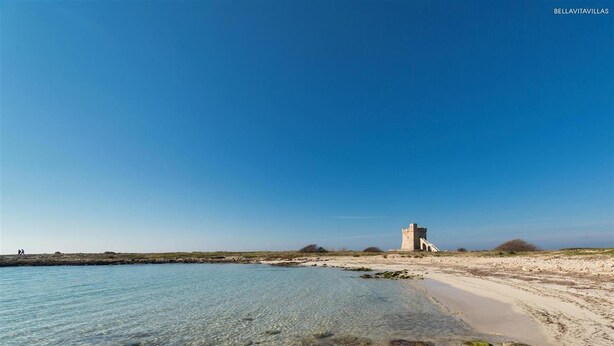 A coastal landscape with clear water, a sandy beach, and a historical stone structure.