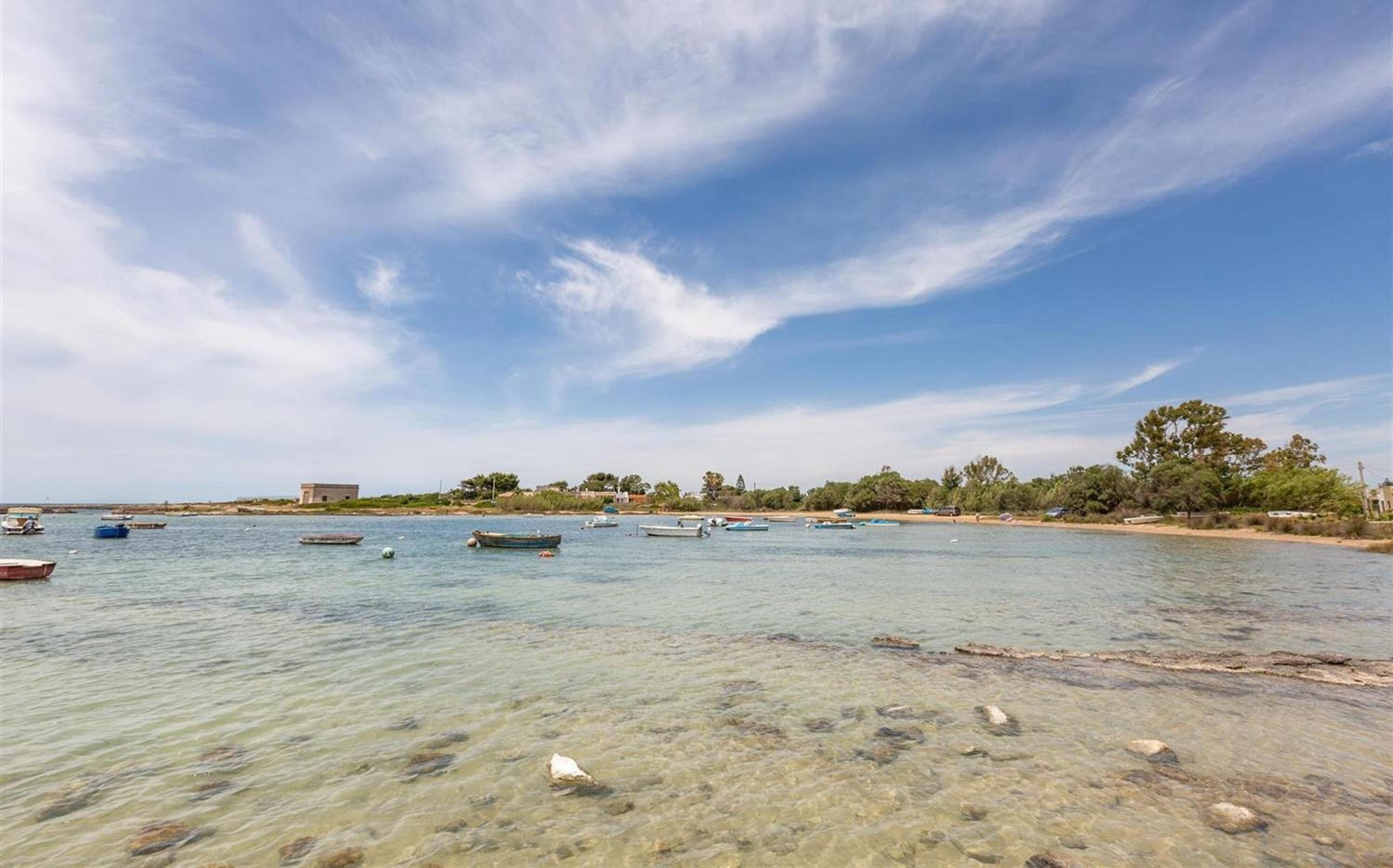 Eine Küstenlandschaft mit Booten auf ruhigem Wasser, blauem Himmel und einem Sandstrand mit vereinzelter Vegetation.