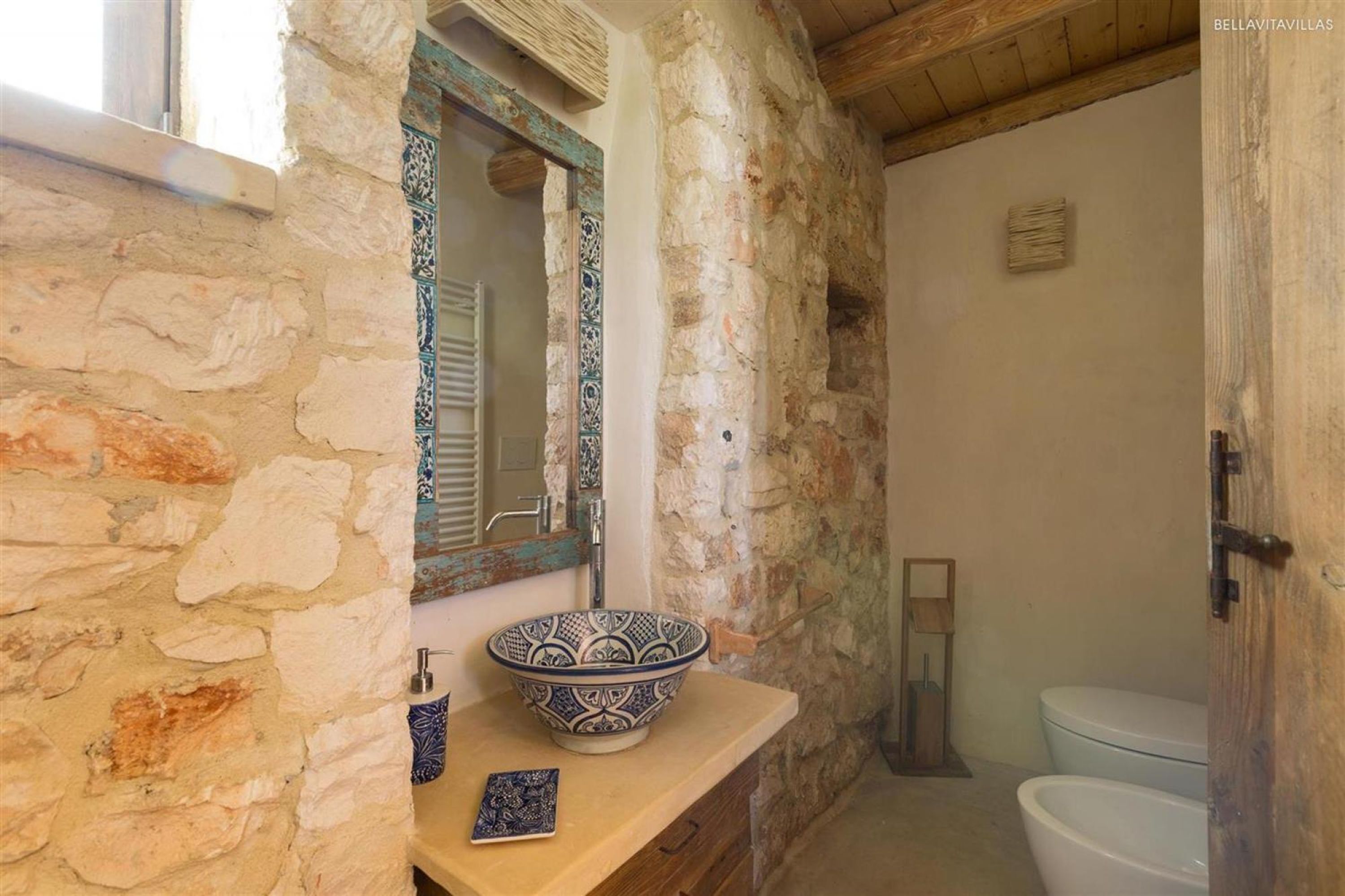 A rustic bathroom with a stone wall, a wooden vanity, a decorative bowl, and a toilet.