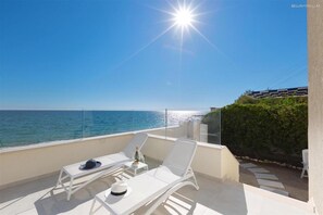 En balkon med to hvide liggestole, et lille bord med en flaske og en hat, og udsigt til havet.