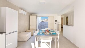 A compact room with a dining table set for four, a sofa, a white refrigerator, and an air conditioning unit. - 1685 Villa Diamante in Campomarino (Maruggio)