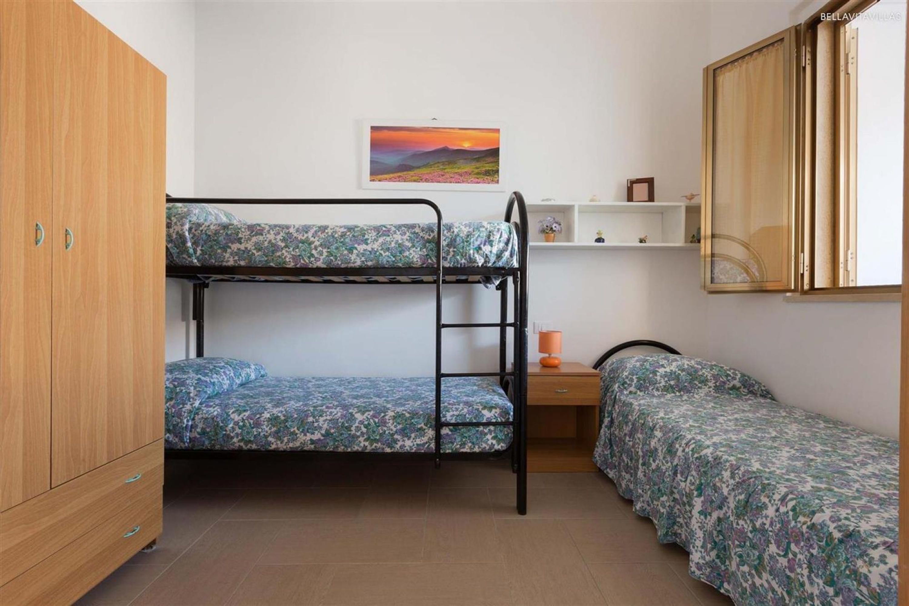 A dormitory room with bunk beds, a wooden wardrobe, a small desk, and a shelf with personal items.